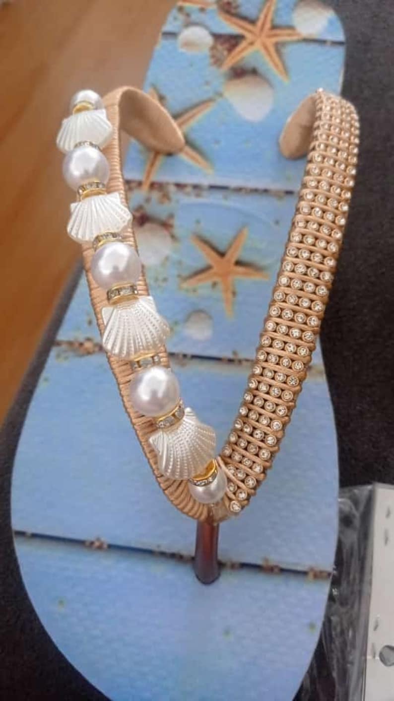 Op de afbeelding: Een paar blauwe slippers met een gouden band versierd met strass steentjes en witte schelpen met parel accenten.