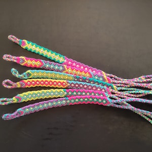 Puede incluir: Un conjunto de pulseras de amistad coloridas con un patrón de chevron y adornos plateados. Las pulseras están hechas con una variedad de colores, incluyendo rosa, amarillo, verde y azul.