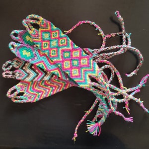 Puede incluir: Una colección de coloridas pulseras de la amistad con intrincados patrones geométricos. Las pulseras presentan una vibrante mezcla de hilos rosas, turquesas, amarillos y blancos, acentuados con pequeños adornos brillantes. Lazos trenzados se extienden de cada pulsera.