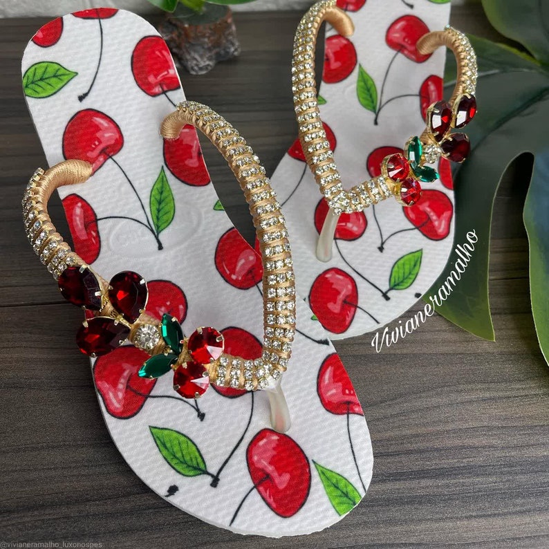 May include: White flip flops with a cherry print and gold and red rhinestone embellishments. The flip flops have a gold strap with rhinestones.