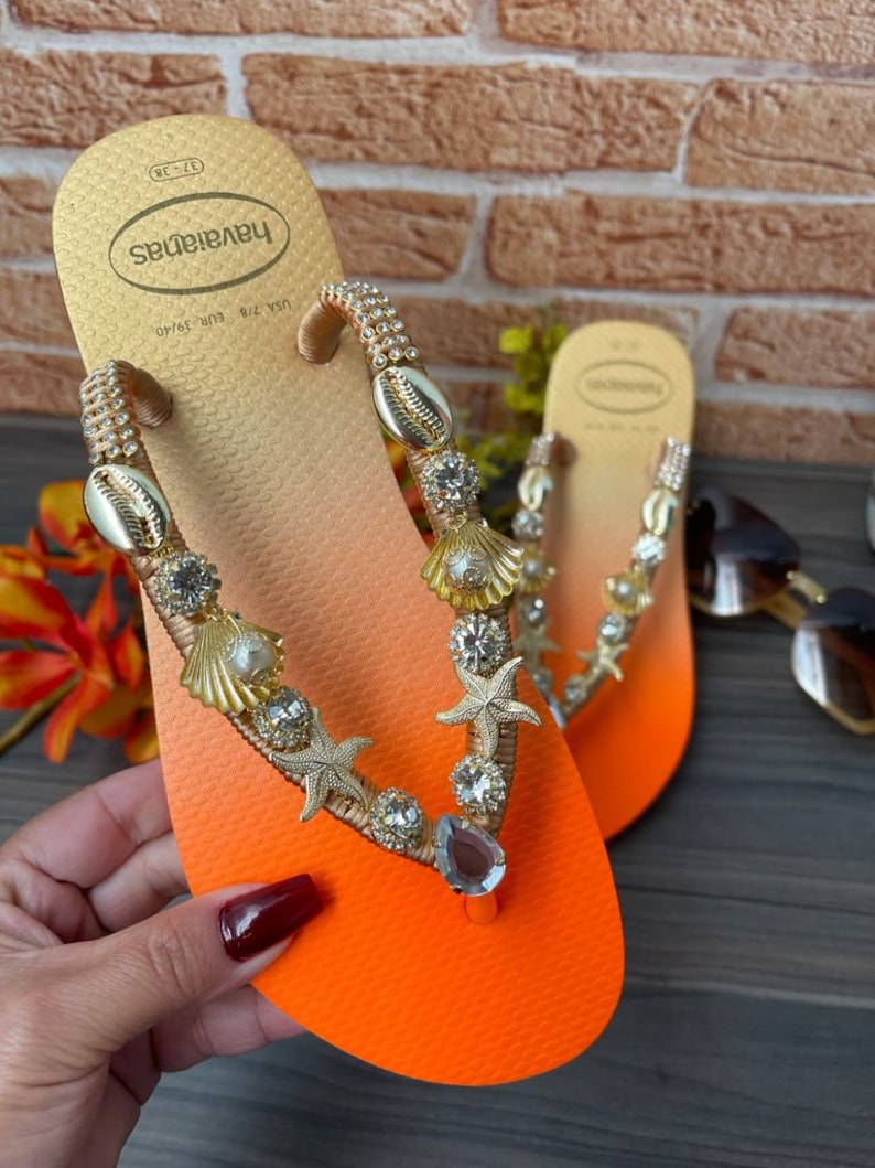 Puede incluir: Chanclas Havaianas naranjas y doradas con correas adornadas. Las correas est&aacute;n adornadas con conchas marinas, estrellas de mar y cristales brillantes. Las chanclas tienen un dise&ntilde;o degradado, que pasa de dorado a naranja.
