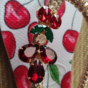 May include: A gold-tone charm with red and green rhinestones. The charm is shaped like a cherry with a green leaf and a red stem.