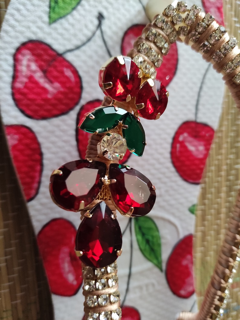 May include: A gold-tone charm with red and green rhinestones. The charm is shaped like a cherry with a green leaf and a red stem.
