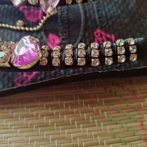May include: Close-up of a black flip-flop with pink rhinestones and a gold chain. The flip-flop has a row of clear rhinestones on a black band.