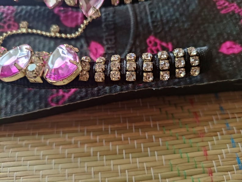 May include: Close-up of a black flip-flop with pink rhinestones and a gold chain. The flip-flop has a row of clear rhinestones on a black band.