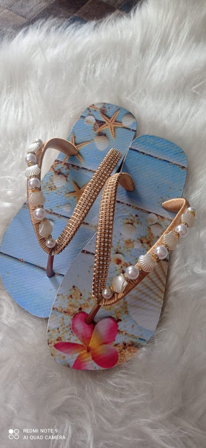 Op de afbeelding: Een paar blauwe slippers met een schelp- en zeesterontwerp. De bandjes zijn versierd met gouden kralen en witte parels.