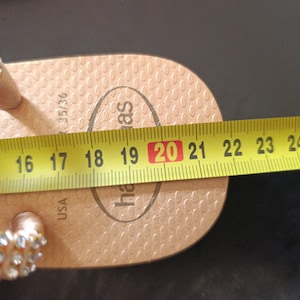 Puede incluir: Una chancla Havaianas de color beige con una correa enjoyada se mide con una cinta m&eacute;trica amarilla. La chancla mide aproximadamente 20 cm de largo. La talla de la chancla es 35/36.