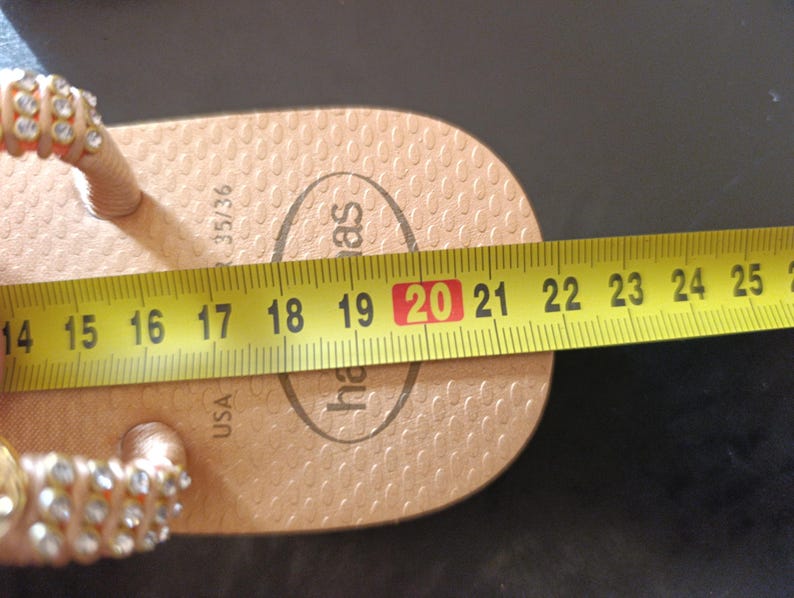 Puede incluir: Una chancla Havaianas de color beige con una correa enjoyada se mide con una cinta m&eacute;trica amarilla. La chancla mide aproximadamente 20 cm de largo. La talla de la chancla es 35/36.