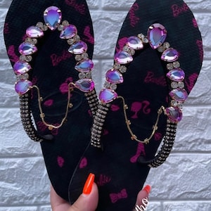 May include: A pair of black flip-flops with pink Barbie logos and rhinestones. The flip-flops have a gold chain detail and iridescent pink gems.