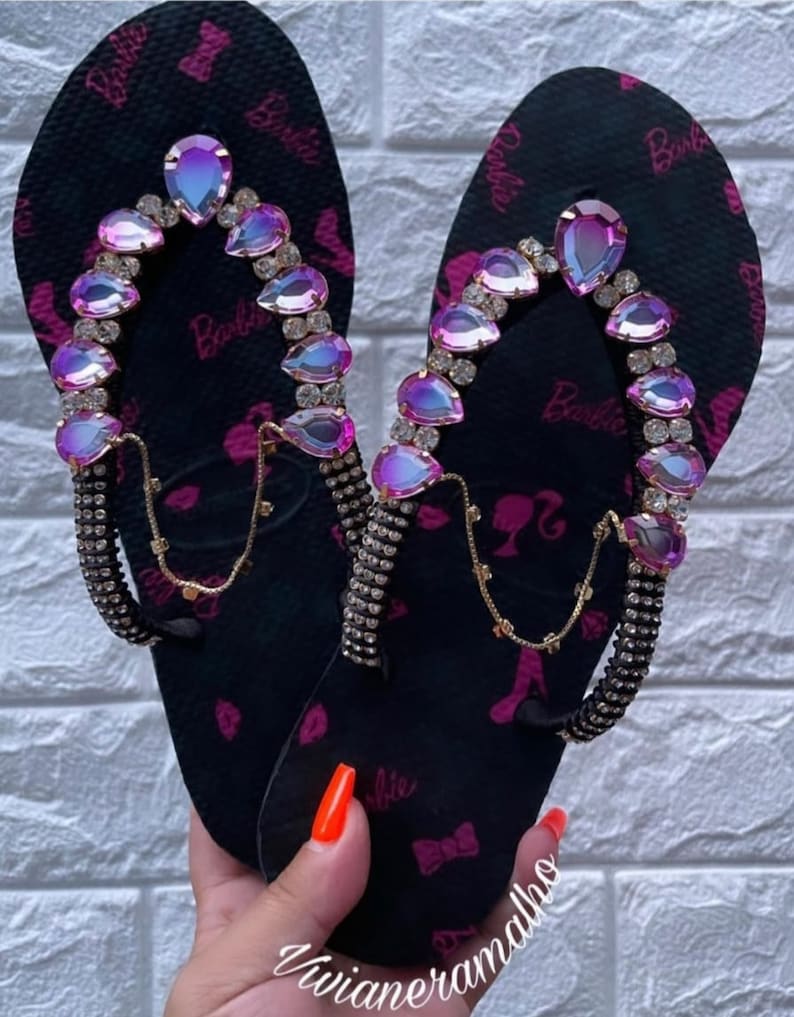 May include: A pair of black flip-flops with pink Barbie logos and rhinestones. The flip-flops have a gold chain detail and iridescent pink gems.