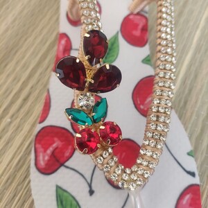May include: A pair of white flip-flops with a cherry print. The flip-flops are decorated with rhinestones and a red, green, and gold cherry charm.