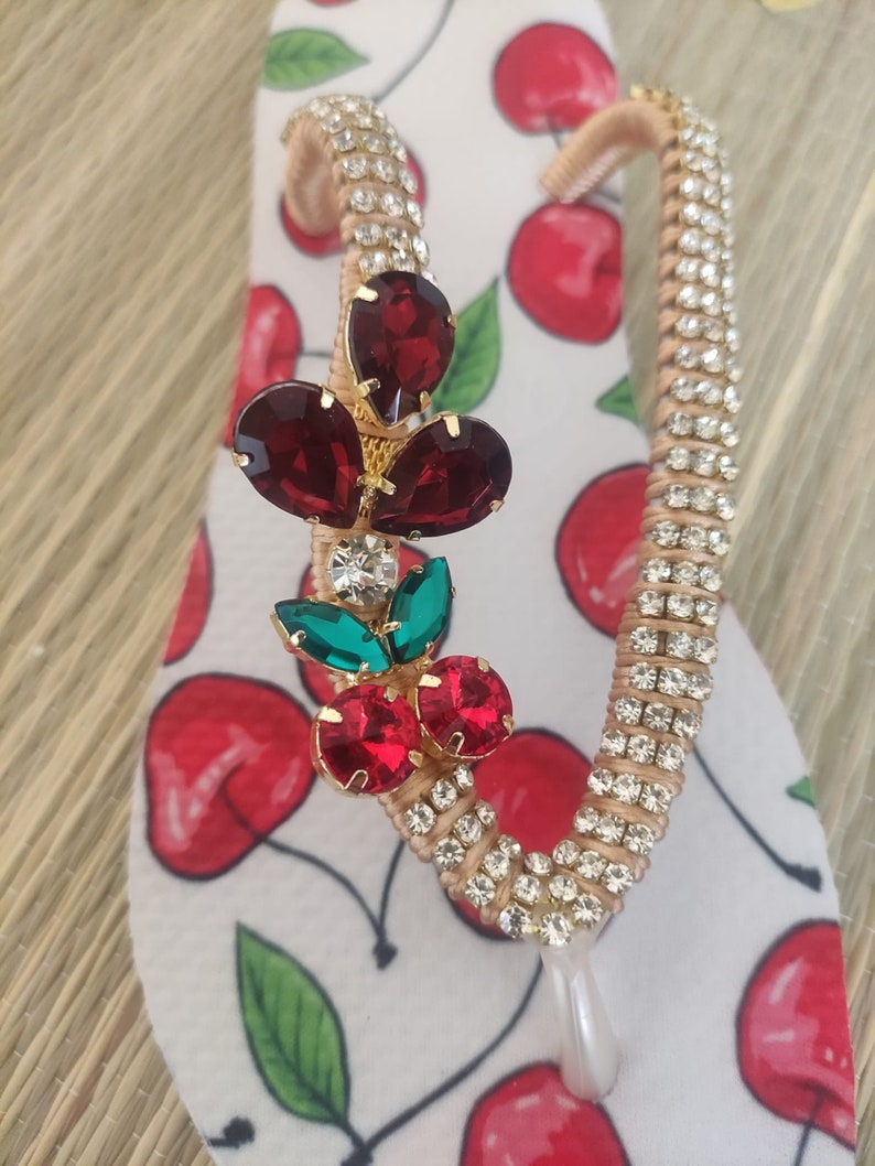 May include: A pair of white flip-flops with a cherry print. The flip-flops are decorated with rhinestones and a red, green, and gold cherry charm.