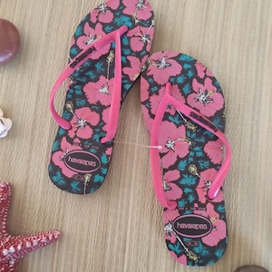 Puede incluir: Un par de chanclas negras con estampado floral rosa. Las chanclas tienen una tira rosa y el nombre de la marca "havaianas" est&aacute; impreso en la suela.