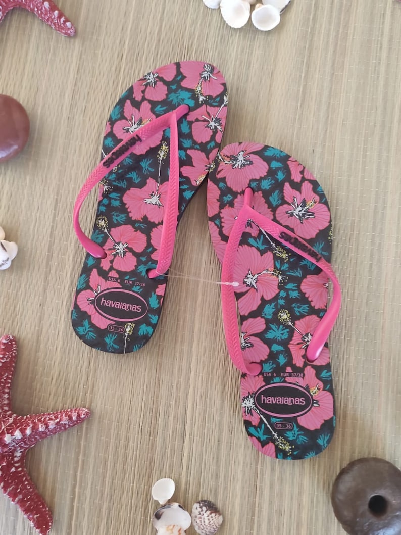 Puede incluir: Un par de chanclas negras con estampado floral rosa. Las chanclas tienen una tira rosa y el nombre de la marca "havaianas" est&aacute; impreso en la suela.