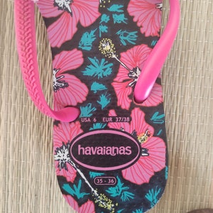 Puede incluir: Chanclas rosa y negras con estampado floral. Las chanclas tienen una tira negra y una suela rosa. Las chanclas son talla 6 USA, 37/38 EUR, 35-36. El nombre de la marca "havaianas" est&aacute; impreso en la suela.