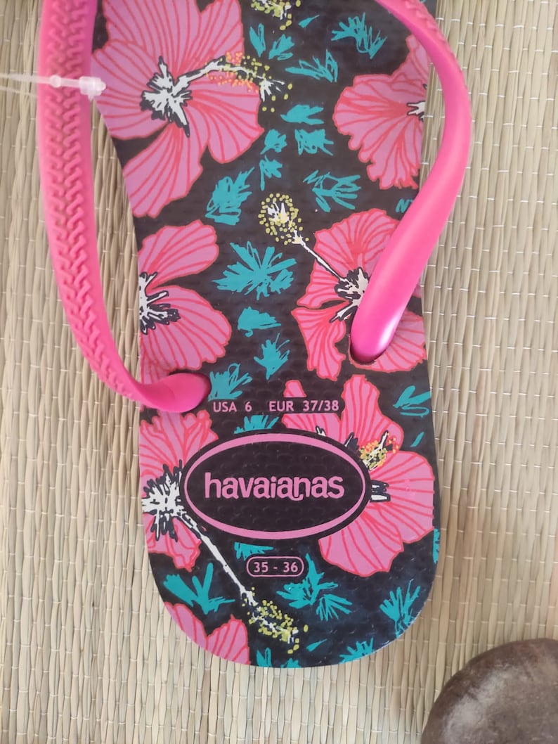 Puede incluir: Chanclas rosa y negras con estampado floral. Las chanclas tienen una tira negra y una suela rosa. Las chanclas son talla 6 USA, 37/38 EUR, 35-36. El nombre de la marca "havaianas" est&aacute; impreso en la suela.