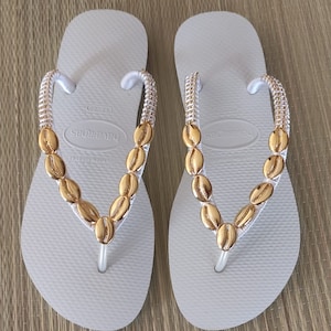 Könnte beinhalten: Weiße Flip-Flops mit einem goldenen Muschel-Design. Die Flip-Flops haben einen weißen Riemen und eine weiße Sohle. Der Markenname "havaianas" ist auf der Sohle aufgedruckt.
