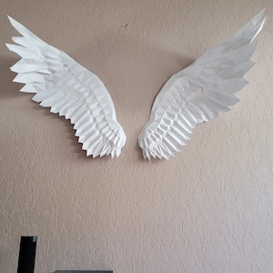 Peut inclure: Une paire d'ailes en papier blanc, grandes et faites de papier plié, sont montées sur un mur rose clair. Les ailes sont symétriques et ont un design géométrique.