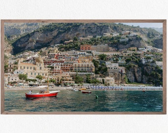 May include: A red boat with a white cabin is docked in the water in front of a colorful town built on a steep cliffside.