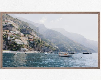 May include: A white boat with a blue stripe is anchored in the water in front of a hillside town. The town is built into the side of a steep, rocky cliff. The water is a deep blue and the sky is a light blue.
