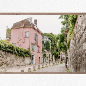 May include: A narrow, cobblestone street in a European city with a pink building on the left and a stone wall on the right. Lush greenery covers the buildings and walls, creating a picturesque scene.