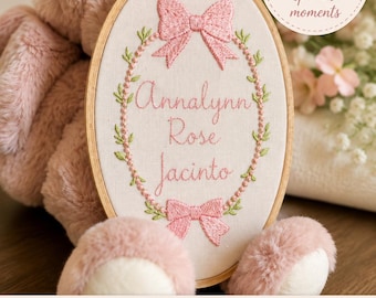 May include: A personalized baby keepsake featuring an embroidered name within a wooden hoop. The design includes pink bows, a floral wreath, and the name "Annalynn Rose Jacinto." The background includes a plush pink teddy bear.