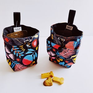 Op de afbeelding: Twee zwarte stoffen zakjes voor hondenkoekjes met een kleurrijk bloemenpatroon. Elk zakje heeft een zwart handvat en een drukknoopsluiting. Er liggen hondenkoekjes naast.