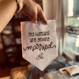 May include: White dog bandana with the text "my humans are getting married" in black script. The bandana has a diamond ring graphic and a small pink heart. The bandana is held up by a hand.