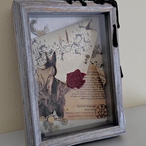 Puede incluir: Una caja de sombra enmarcada con un mapa vintage, un sello de cera y una pequeña figura de metal. El marco es de madera desgastada y tiene un acabado desgastado. La caja de sombra está decorada con cera negra que gotea desde la parte superior.