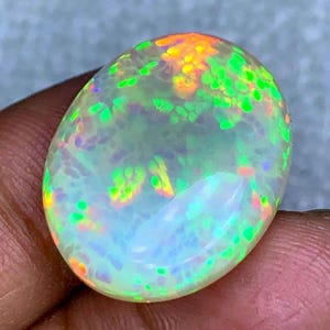 May include: An oval-shaped opal gemstone with a translucent, milky white base. The opal displays vibrant flashes of color, including green, orange, and yellow, creating a play-of-color effect. The gemstone is held in a person's hand.