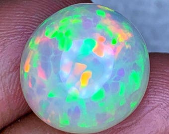May include: A round, cabochon-cut opal gemstone featuring a translucent white base with vibrant, multi-colored flashes of neon green, bright orange, and soft purple throughout its surface, displaying a high-domed, polished, and reflective finish.