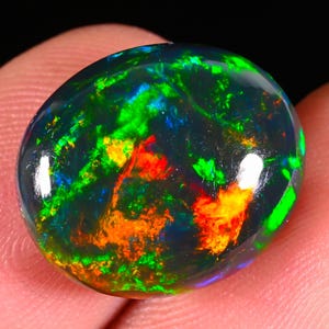 Puede incluir: Una piedra preciosa de ópalo negro pulido de forma ovalada. El ópalo muestra colores internos vibrantes, incluyendo verde, naranja, rojo y azul, creando una apariencia dinámica e iridiscente. La piedra preciosa se sostiene entre los dedos.