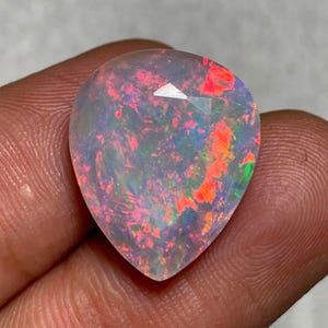 May include: A pear-shaped opal gemstone featuring a translucent body with vibrant flashes of red, orange, and green play-of-color. The stone has a faceted surface that reflects light, showcasing a distinct internal pattern of bright neon hues.