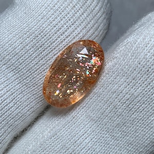 Può includere: Una pietra preziosa di pietra del sole di forma ovale con superficie sfaccettata, che mostra una tonalità arancione traslucida e inclusioni metalliche interne color rame che riflettono la luce con scintillii rosa, verdi e dorati.