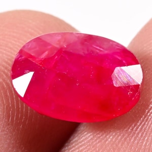 Peut inclure: Une pierre précieuse de type rubis, taillée en forme ovale. La gemme est d'un rouge profond et vibrant, avec une surface polie. Le rubis est tenu entre deux doigts, mettant en valeur sa taille et sa brillance.