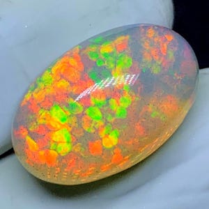 May include: An oval-shaped opal gemstone with a vibrant display of color. The opal features a mix of orange, yellow, and green hues, with a shimmering, iridescent effect. The gemstone is set against a neutral background, highlighting its natural beauty.