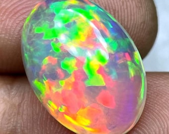 May include: An oval-shaped opal gemstone with a vibrant display of colors. The stone features a mix of green, yellow, orange, and pink hues, creating a dynamic and iridescent effect. The opal is held between fingers, showcasing its polished surface.