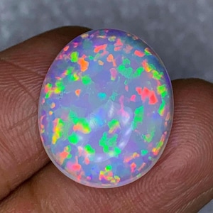 Puede incluir: Una piedra preciosa en forma de cabujón ovalado con una base blanca translúcida y un vibrante juego de colores que presenta motas dispersas de verde neón brillante, naranja y rosa en toda la superficie, creando un efecto iridiscente luminoso.