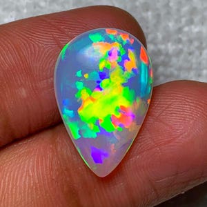Könnte beinhalten: Ein birnenförmiger Opal-Edelstein mit einer durchscheinenden weißen Basis und lebhaften Blitzen in Neongrün, hellem Orange und tiefem Violett auf der Oberfläche. Der Stein hat einen glatten, polierten Cabochon-Schliff mit abgerundeter Oberseite.