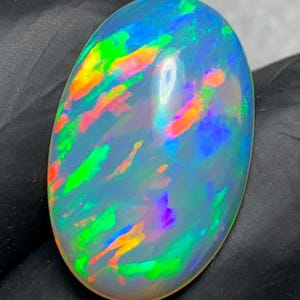 May include: An oval-shaped cabochon opal gemstone featuring a translucent white base with vibrant flashes of neon green, electric blue, and bright orange color play distributed across the smooth, polished, and domed surface of the stone.