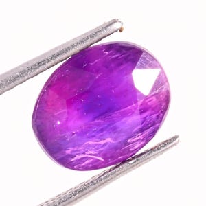 May include: A faceted, oval-shaped gemstone with a gradient of purple and pink hues. The gem is held by silver tweezers against a white background, showcasing its polished surfaces and internal color variations. The gem's facets reflect light.
