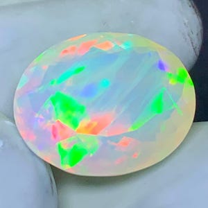 May include: A round, faceted opal gemstone with a milky white base color. The opal displays vibrant flashes of green, orange, pink, and blue, creating a play-of-color effect. The gemstone is set against a neutral background.