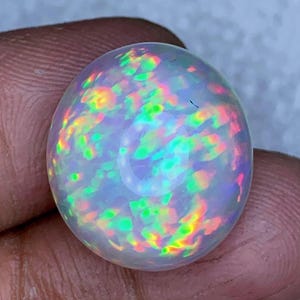 May include: A polished, oval-shaped opal gemstone. The opal displays a milky white base color with vibrant flashes of iridescent colors, including green, yellow, orange, and pink. The gemstone is held between two fingers, showcasing its natural beauty.