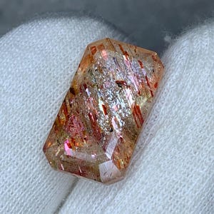 Può includere: Una pietra preziosa di pietra del sole a taglio rettangolare sfaccettato, caratterizzata da una base traslucida color pesca e arancio con distintive inclusioni metalliche bruno-rossastre e riflessi scintillanti interni in tutta la struttura cristallina.