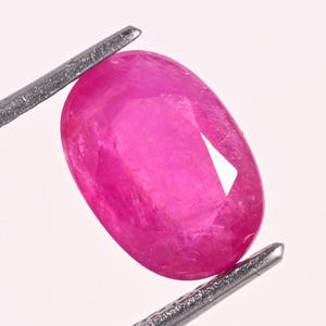 Puede incluir: Una gema de rubí facetada, de forma ovalada, sujeta con pinzas metálicas. El rubí es de un color rojo-rosado vibrante, con inclusiones internas visibles y una superficie pulida. Las facetas de la gema capturan la luz.