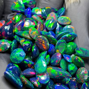 May include: A collection of faceted black opals featuring vibrant flashes of neon green, electric blue, and fiery red color play. Each gemstone is individually cut with multiple polished surfaces that reflect light, creating a brilliant display.