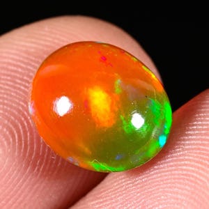 May include: A polished oval-shaped fire opal gemstone featuring a vibrant orange base color with internal flashes of bright green, yellow, and red light. The gemstone has a smooth, rounded cabochon surface that reflects light across its surface.