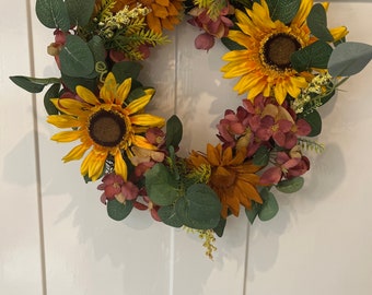 May include: A sunflower wreath with yellow and orange sunflowers, green leaves, and burgundy flowers.
