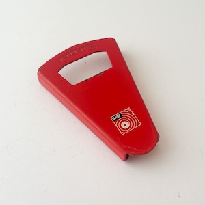 May include: A red bottle opener with a rectangular body and a handle. The opener has a small square logo with concentric circles and the word "BASF". The word "BASF" is also printed on the handle.