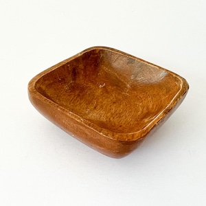 May include: A small, square wooden bowl with rounded corners. The bowl is made of a light brown wood with visible grain and a smooth, polished finish. The interior is slightly darker than the exterior.